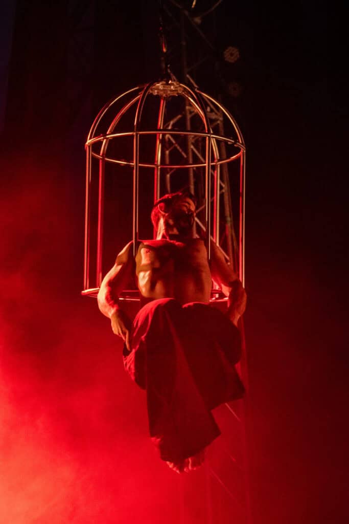Performer inside a birdcage on stage with red lighting and smoke effects.