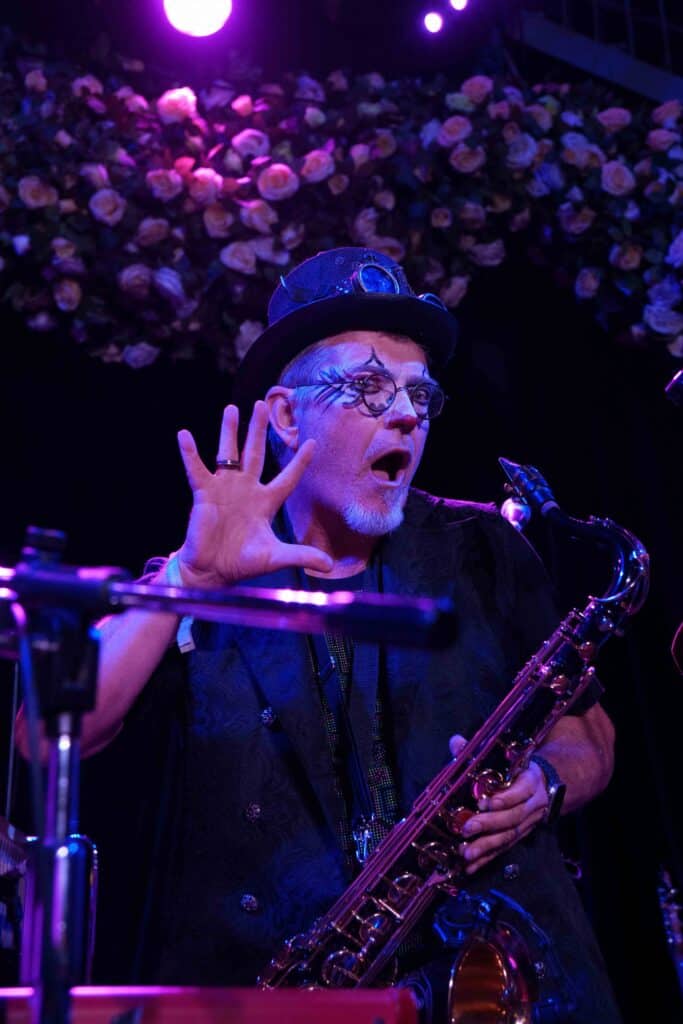 Musician playing saxophone on stage with purple lighting and floral backdrop.