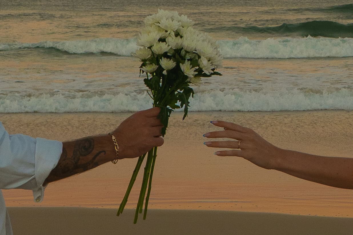 Person handing bouquet of white flowers to another on beach at sunset.